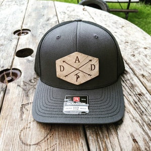 K&ouml;nnte beinhalten: Ein dunkelgrauer Trucker-Hut mit schwarzem Netzr&uuml;cken. Der Hut hat einen Holz-Patch mit den Buchstaben "A D D" und gekreuzten Pfeilen. Der Patch hat eine sechseckige Form. Der Hut hat ein schwarzes Etikett mit dem Text "EST. 1970 RICHARDSON ORIGINAL 112 TRUCKER ADJUSTABLE".