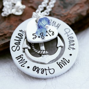 May include: A silver pendant necklace with a nautical theme. The pendant has a circular base with the words "My Sailor, My Hero, My Heart" and an anchor design. A heart-shaped charm with the date "5-8-15" is attached, along with a blue crystal bead.