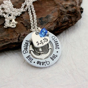 May include: Silver necklace with a pendant featuring an anchor, heart, and the words "My Sailor, My Heart." The heart has the date "5.8.15." A blue crystal bead hangs above the heart. The necklace is on a textured brown surface.