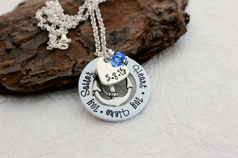 May include: Silver necklace with a pendant featuring an anchor, heart, and the words "My Sailor, My Heart." The heart has the date "5.8.15." A blue crystal bead hangs above the heart. The necklace is on a textured brown surface.