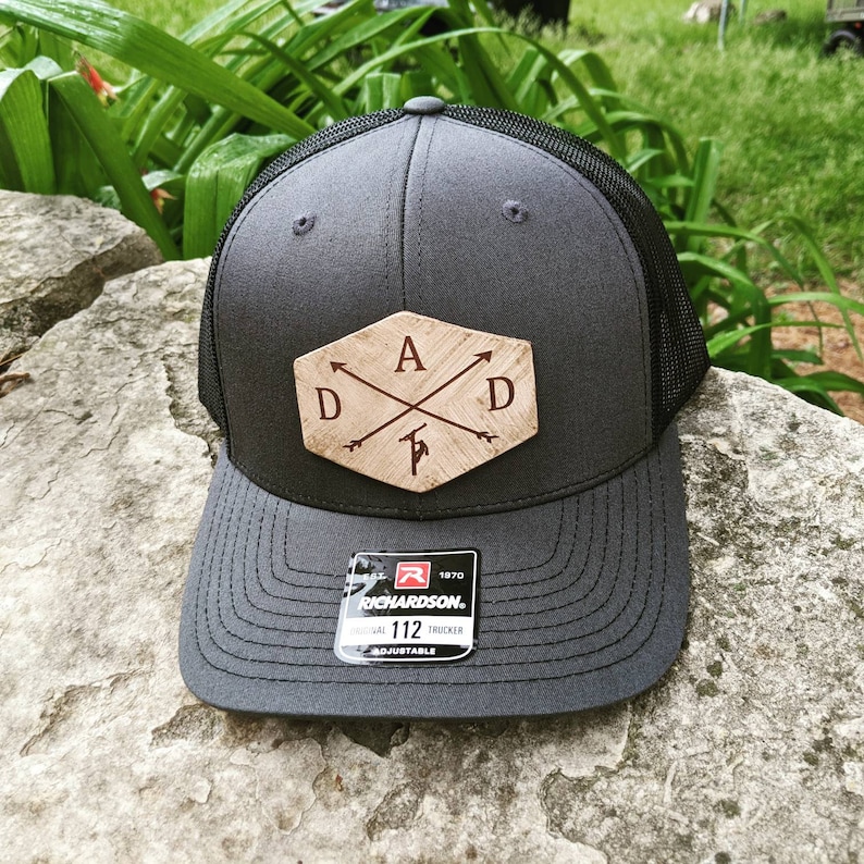 K&ouml;nnte beinhalten: Eine graue und schwarze Trucker-Kappe mit einem braunen Holz-Patch, auf dem "A D" mit Pfeilen steht, die auf die Buchstaben zeigen. Der Patch hat die Form eines Sechsecks. Die Kappe hat ein schwarzes Netzr&uuml;cken und einen schwarzen verstellbaren Riemen mit einem roten "Richardson"-Etikett.