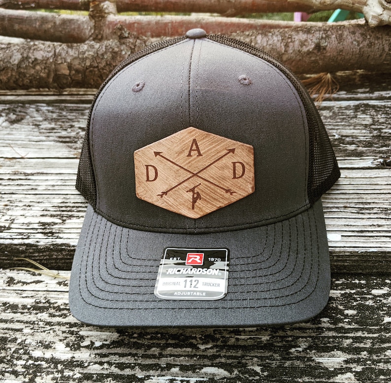 K&ouml;nnte beinhalten: Eine schwarze und graue Trucker-Kappe mit einem Holz-Patch, auf dem die Buchstaben "DAD" mit Pfeilen und einem kleinen Baumdesign zu sehen sind. Der Patch hat eine sechseckige Form. Die Kappe hat ein schwarzes Netzr&uuml;cken und einen schwarzen verstellbaren Riemen mit dem Text "Richardson" und "112" auf einem roten Etikett.
