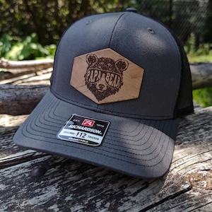Peut inclure: Un chapeau de camionneur gris et noir avec un patch en bois qui dit "Papa Bear" et représente un ours portant des lunettes de soleil. Le chapeau est réglable et porte le logo Richardson 112.