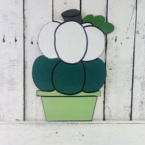 May include: A white pumpkin with a green stem and leaves, painted in a cartoon style, sitting in a green pot. The pumpkin is made of wood and is a fall decoration.