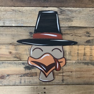 May include: A wooden turkey head cutout with a black pilgrim hat, brown eyes, and an orange beak. The turkey head is painted in shades of brown, orange, and white.