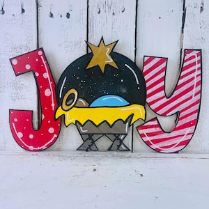 May include: A wooden Christmas sign with the word "JOY" painted in red, white, and black. The "O" is a manger scene with a baby Jesus, a star, and a blue sky.