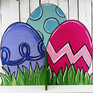 May include: Three large wooden Easter eggs painted in different colors and patterns. The eggs are standing in a bed of green grass. The purple egg has a blue swirl design, the blue egg has white polka dots, and the pink egg has a white zig-zag pattern.