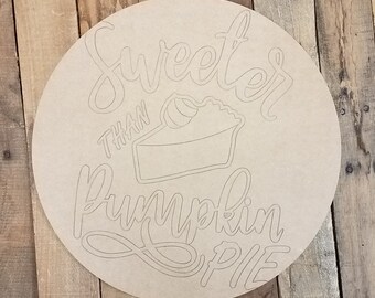 May include: A round wooden sign with the text "Sweeter than Pumpkin Pie" in a handwritten font. A pie slice with a swirl of whipped cream is pictured in the center of the sign.