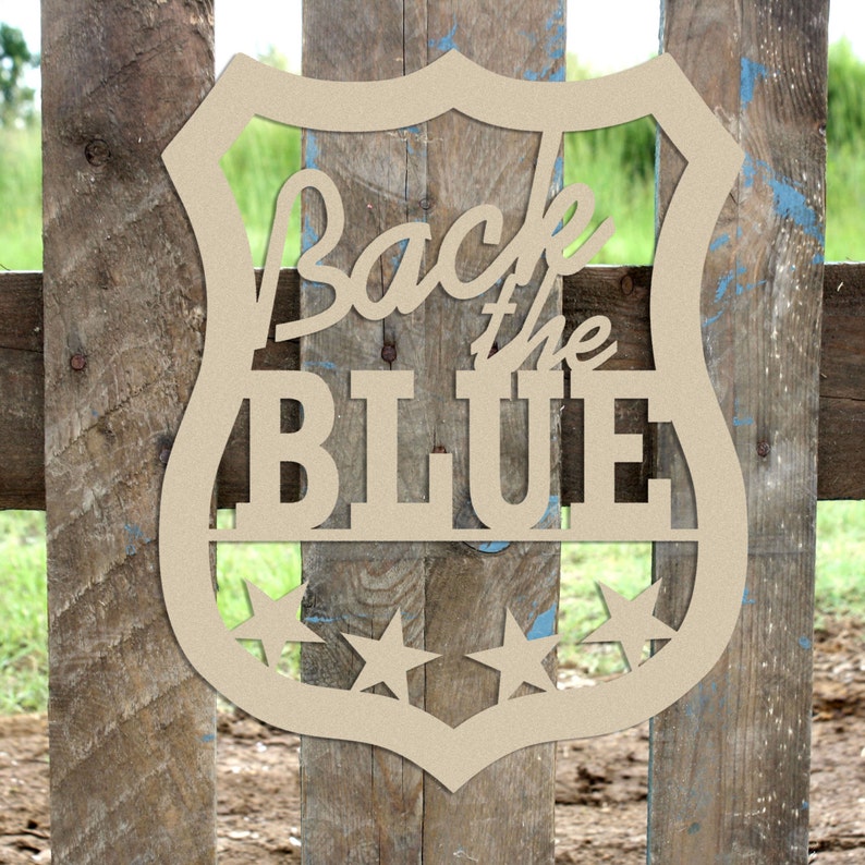 May include: Wooden cutout of a police badge with the text "Back the Blue" and three stars. The badge is brown and has a rounded shape.