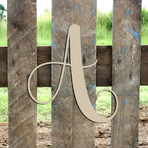 May include: A large, wooden, cursive letter A, painted in a light brown color, is leaning against a weathered wooden fence. The letter is in a decorative script font.