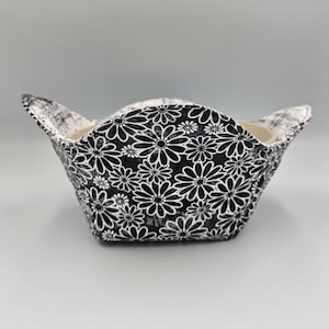 May include: A black and white floral print bowl cozy. The cozy is made of fabric and has a black daisy pattern on a white background. The bowl cozy is designed to hold a bowl and keep the contents warm.