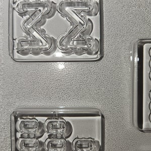 May include: Clear, rectangular molds with various shapes. One mold features two mirrored "Z" shapes, while another contains multiple rounded rectangular forms. The molds are on a textured, gray surface.