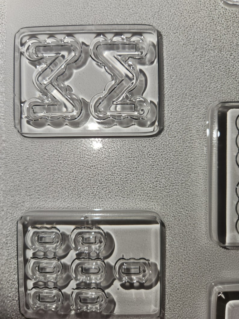 May include: Clear, rectangular molds with various shapes. One mold features two mirrored "Z" shapes, while another contains multiple rounded rectangular forms. The molds are on a textured, gray surface.