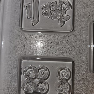 May include: Clear, rectangular glass molds with embossed designs. One mold features Chinese characters, while another has a pattern of six circular shapes. The molds are set against a textured, gray background.