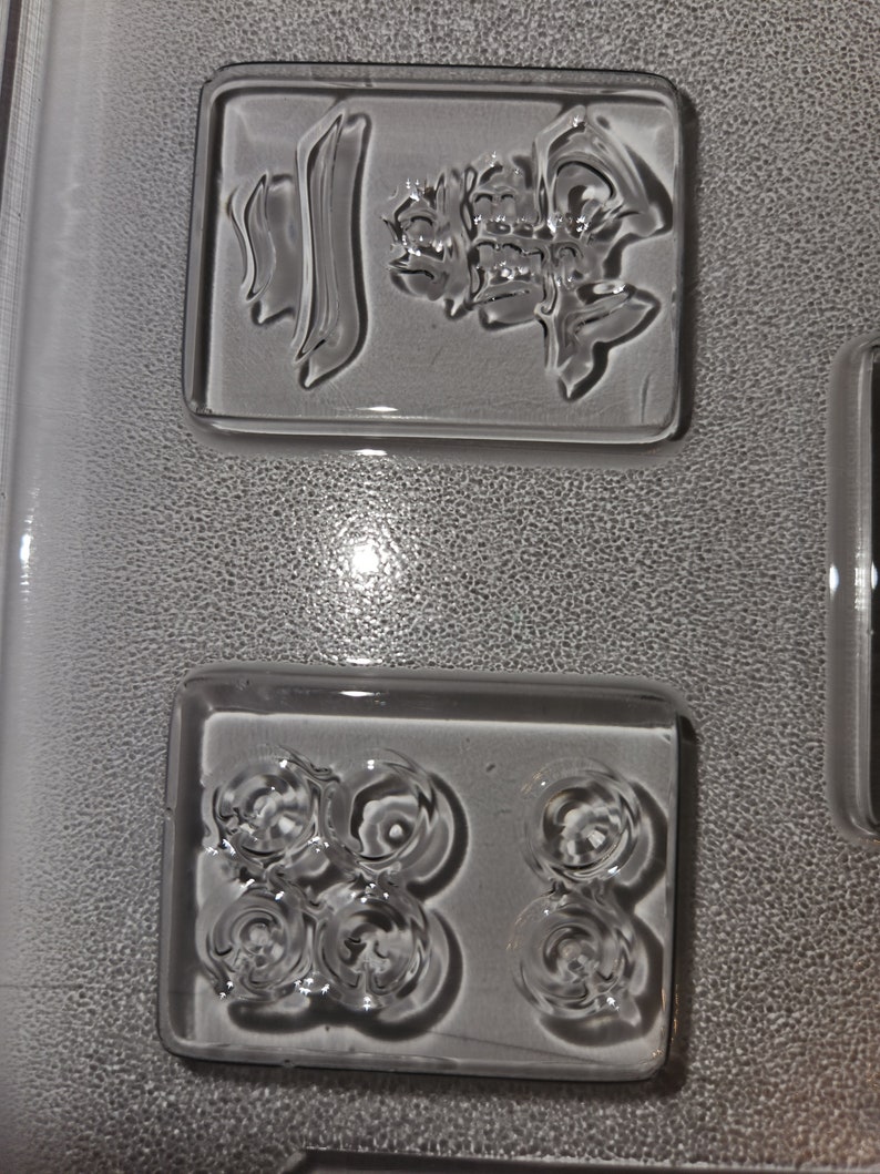May include: Clear, rectangular glass molds with embossed designs. One mold features Chinese characters, while another has a pattern of six circular shapes. The molds are set against a textured, gray background.