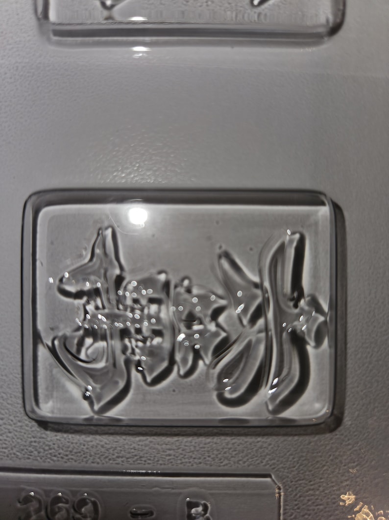 May include: A clear, rectangular mold with embossed Japanese characters. The characters are in a stylized font and appear to be for a food item. The mold is made of a clear, glossy material.