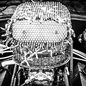 May include: A black and white photo of a car engine with a decorative, silver,  circular object on top. The object is made of metal and has a honeycomb pattern.