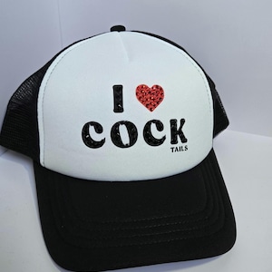 May include: Black and white trucker hat with the phrase "I ❤️ COCK" in black letters and a red heart. The hat has a black brim and mesh back, with the text "TAILS" below the word "COCK".
