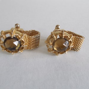 Pu&ograve; includere: Un paio di gemelli in oro con una pietra di vetro sfaccettata marrone incastonata in una cornice decorativa e ornata. I gemelli sono collegati da una catena d'oro.