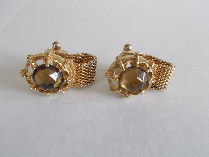 Pu&ograve; includere: Un paio di gemelli in oro con una pietra di vetro sfaccettata marrone incastonata in una cornice decorativa e ornata. I gemelli sono collegati da una catena d'oro.