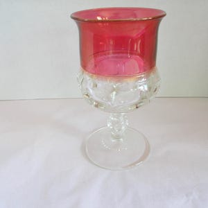May include: A glass goblet featuring a deep red upper bowl and a clear, textured lower section with a repeating embossed pattern. The stem is clear glass with a small decorative node, leading down to a wide, circular, clear glass base.