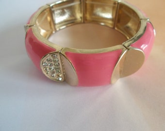 May include: A pink and gold bangle bracelet with a row of clear rhinestones. The bracelet is made of metal and has a smooth, glossy finish.