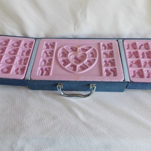 May include: A blue, fabric-covered case with a silver handle, open to reveal a pink plastic mold with heart-shaped and other geometric designs.