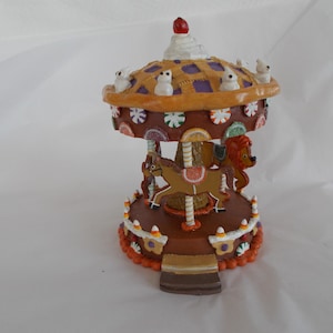 May include: A decorative carousel figurine with a brown base and a multi-tiered design. The top features a gingerbread-style roof with white accents and small animal figures. The carousel has a horse and lion figure, with colorful details and a festive appearance.