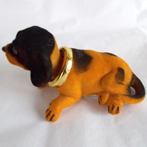 May include: A vintage dog figurine with a black head and spots on its orange body. It has a gold-colored collar. The dog is made of a soft, velvety material.