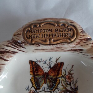 May include: A white ceramic dish with a brown and white woodgrain design. The dish has a raised inscription that reads "Hampton Beach New Hampshire". A butterfly with brown, orange, and blue wings is painted on the dish.