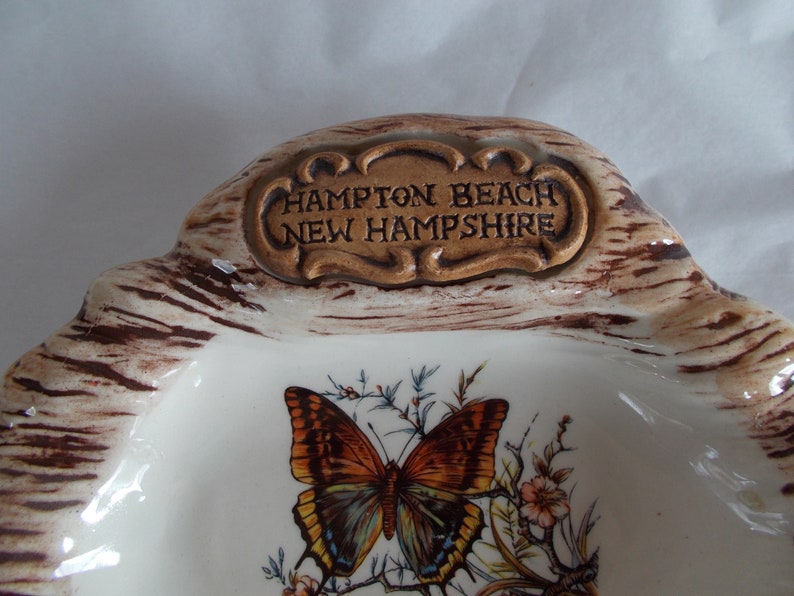 May include: A white ceramic dish with a brown and white woodgrain design. The dish has a raised inscription that reads "Hampton Beach New Hampshire". A butterfly with brown, orange, and blue wings is painted on the dish.