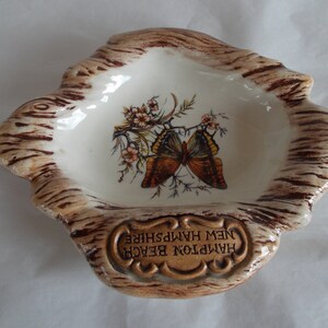 May include: A ceramic ashtray shaped like a tree stump with a brown and white glaze. The ashtray features a painted butterfly and flowers. The text "Hampton Beach New Hampshire" is inscribed on the front.