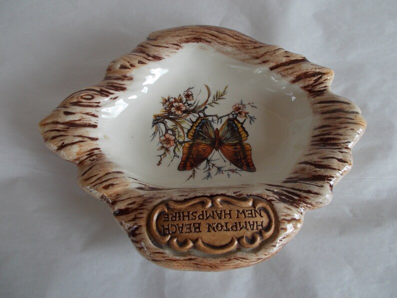 May include: A ceramic ashtray shaped like a tree stump with a brown and white glaze. The ashtray features a painted butterfly and flowers. The text "Hampton Beach New Hampshire" is inscribed on the front.