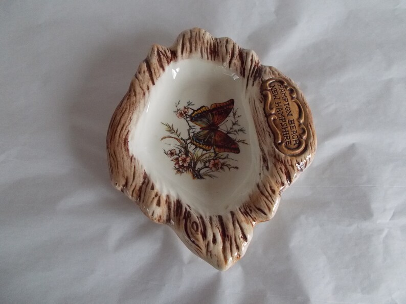 May include: A small, white ceramic dish shaped like a tree stump with a brown butterfly design and the text "Hampton Beach New Hampshire".