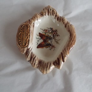 May include: A small, white ceramic dish shaped like a tree stump with a brown glaze. The dish features a painted butterfly and the text "Hampton Beach, New Hampshire".