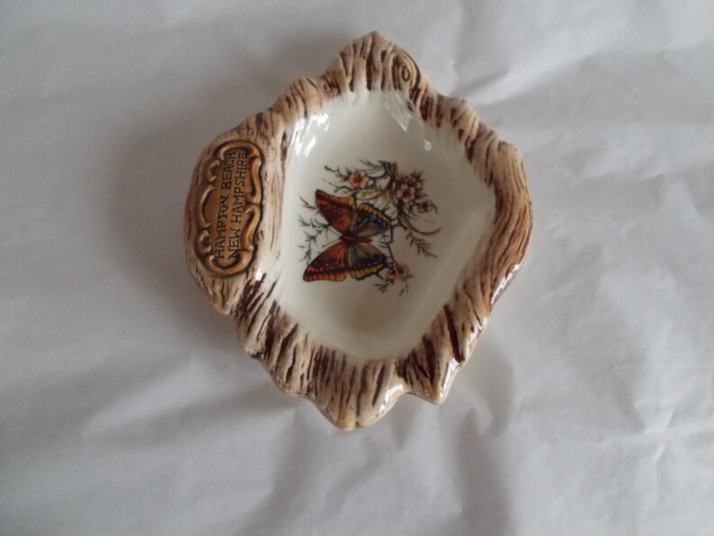 May include: A small, white ceramic dish shaped like a tree stump with a brown glaze. The dish features a painted butterfly and the text "Hampton Beach, New Hampshire".