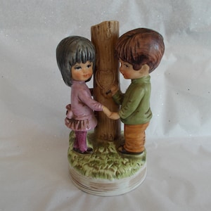 May include: A porcelain figurine of a girl and boy holding hands in front of a tree with a heart carved into it. The figurine is painted in shades of brown, green, pink, and orange.