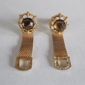 Pu&ograve; includere: Un paio di gemelli d'oro con una pietra di vetro marrone al centro di ciascuno. I gemelli hanno un design a catena e una chiusura a forma di D.