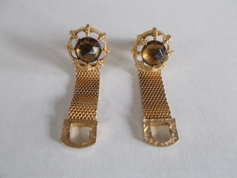 Pu&ograve; includere: Un paio di gemelli d'oro con una pietra di vetro marrone al centro di ciascuno. I gemelli hanno un design a catena e una chiusura a forma di D.