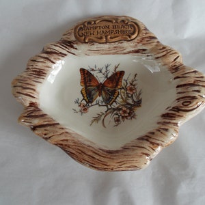 May include: A ceramic ashtray shaped like a tree stump with a brown and white woodgrain design. The ashtray features a butterfly with orange and brown wings and a white background. The text "Hampton Beach New Hampshire" is written on the top of the ashtray.
