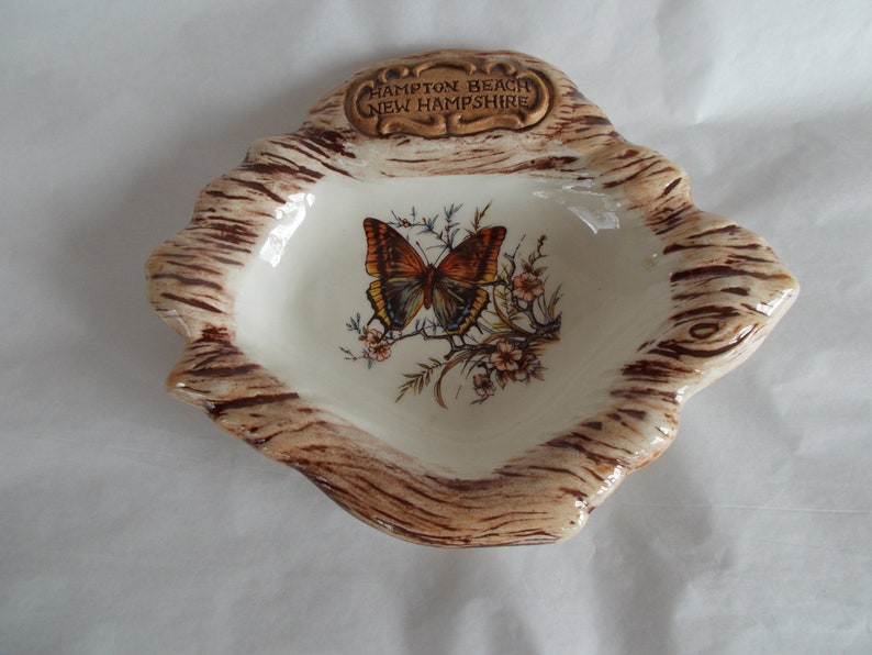 May include: A ceramic ashtray shaped like a tree stump with a brown and white woodgrain design. The ashtray features a butterfly with orange and brown wings and a white background. The text "Hampton Beach New Hampshire" is written on the top of the ashtray.