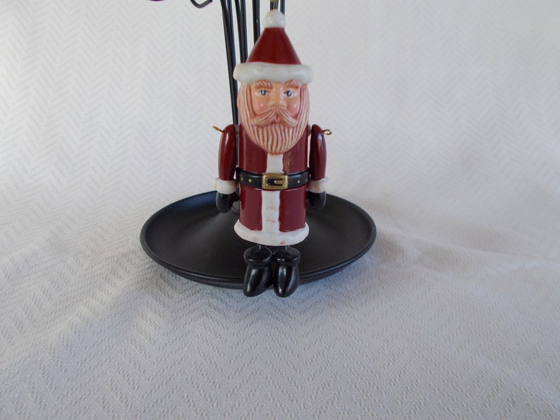 Op de afbeelding: Een rood en wit keramiek kerstman ornament met een zwarte riem en zwarte laarzen. Het ornament hangt aan een zwarte metalen standaard.