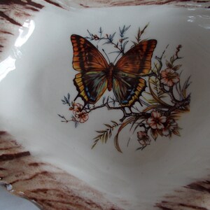 May include: A white ceramic dish with a brown woodgrain pattern around the edge. The center of the dish features a painted butterfly with brown, orange, and blue wings, surrounded by green and yellow foliage and white flowers.