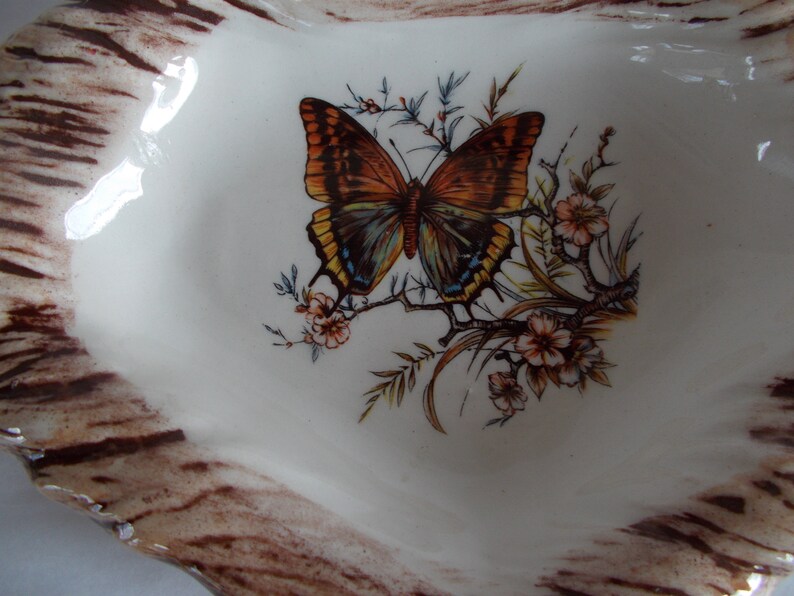 May include: A white ceramic dish with a brown woodgrain pattern around the edge. The center of the dish features a painted butterfly with brown, orange, and blue wings, surrounded by green and yellow foliage and white flowers.