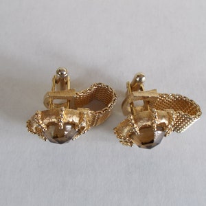 Pu&ograve; includere: Un paio di gemelli d'oro con una pietra marrone al centro. I gemelli hanno un design a catena.