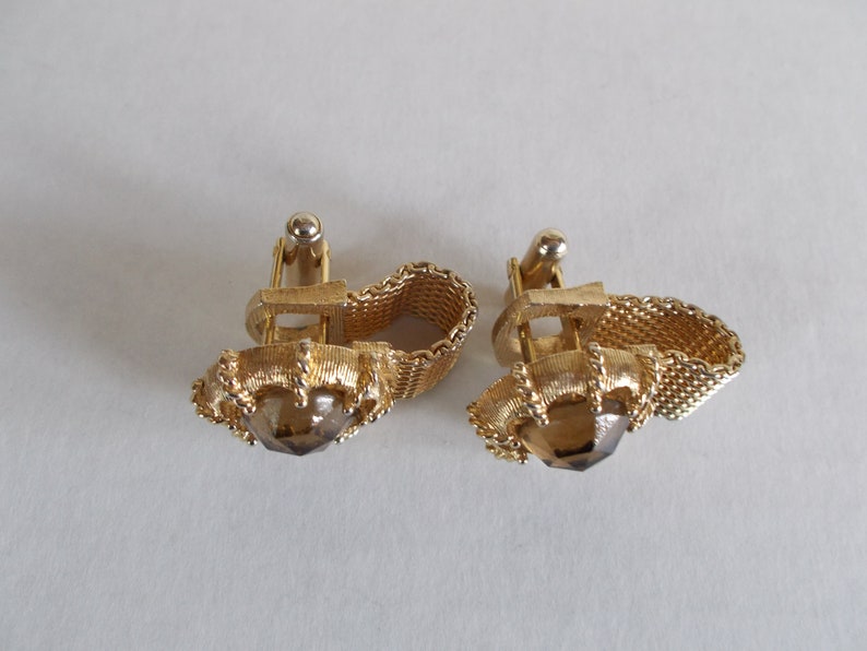 Pu&ograve; includere: Un paio di gemelli d'oro con una pietra marrone al centro. I gemelli hanno un design a catena.