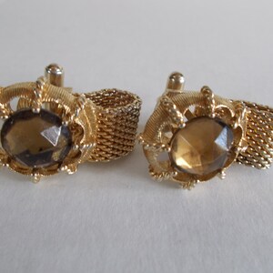 Pu&ograve; includere: Un paio di gemelli d'oro con una pietra di vetro sfaccettata marrone al centro di ogni gemello. I gemelli sono collegati da una catena d'oro.