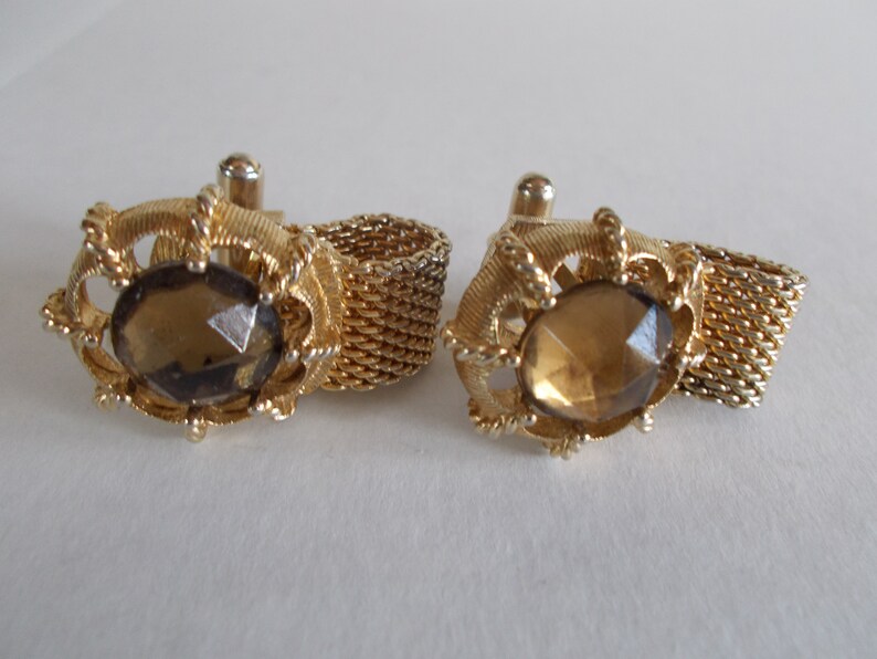 Pu&ograve; includere: Un paio di gemelli d'oro con una pietra di vetro sfaccettata marrone al centro di ogni gemello. I gemelli sono collegati da una catena d'oro.