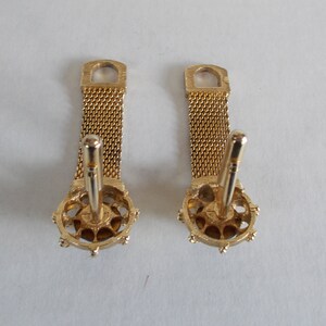 Pu&ograve; includere: Un paio di gemelli d'oro con una catena a maglia e un design rotondo e ornato. I gemelli hanno una finitura dorata e sono in buone condizioni.