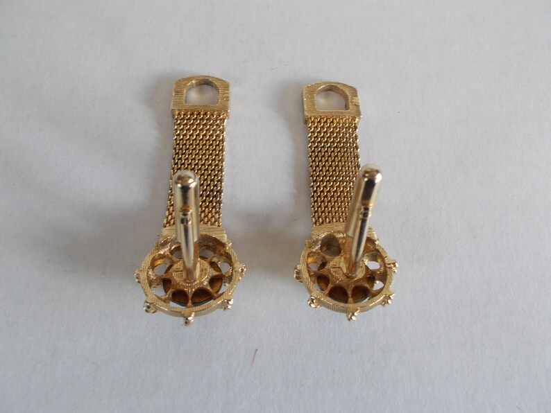 Pu&ograve; includere: Un paio di gemelli d'oro con una catena a maglia e un design rotondo e ornato. I gemelli hanno una finitura dorata e sono in buone condizioni.